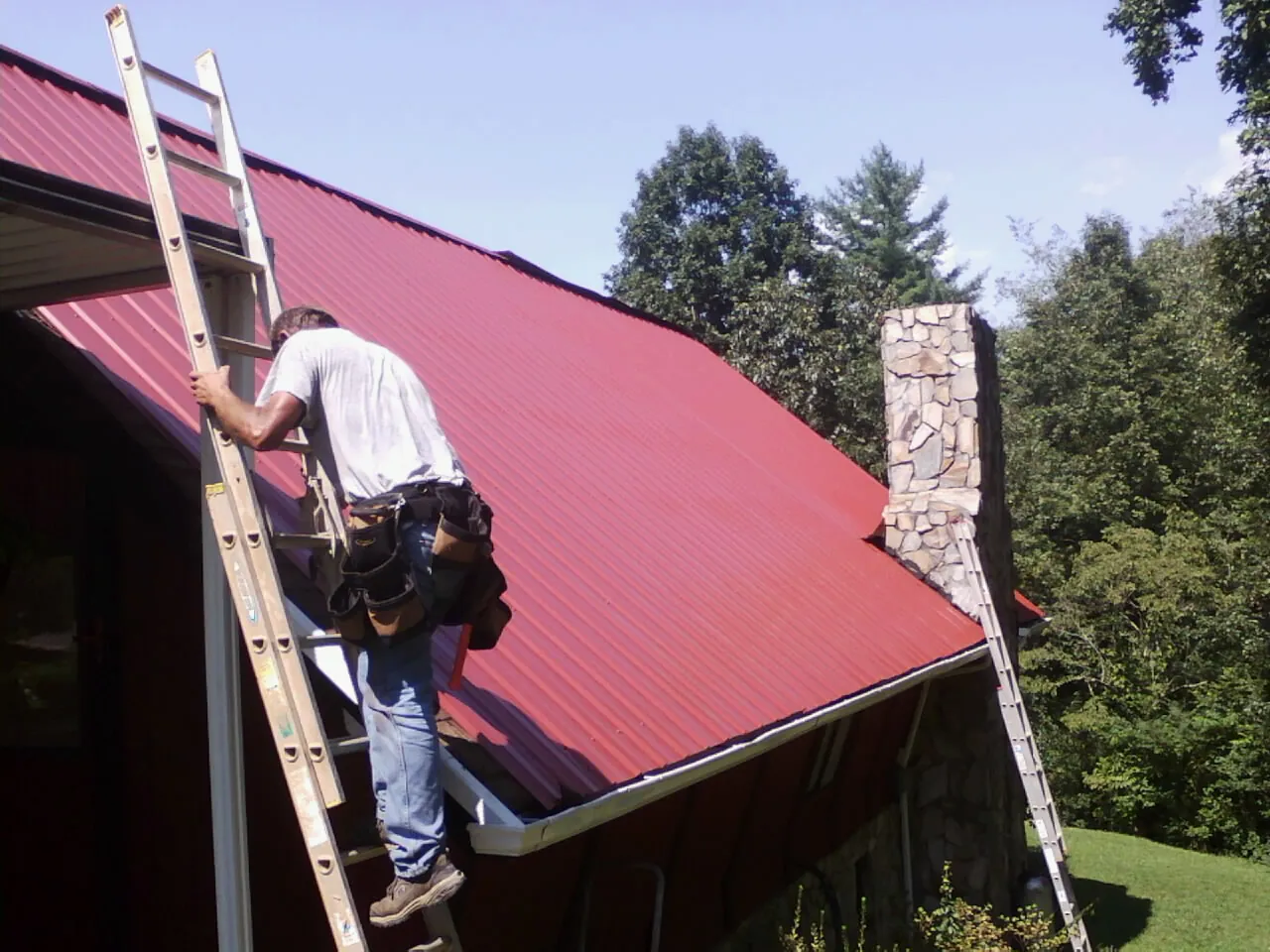 Roofer on a ladder beside a red metal roof and stone chimney — Soffit & Fascia Repair by Roofing Brothers in Willseyville